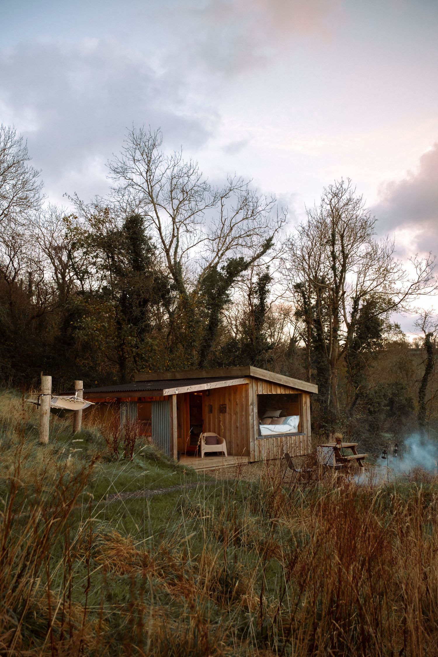 Rustic wooden cabin surrounded by tall grass and bare trees at sunset.