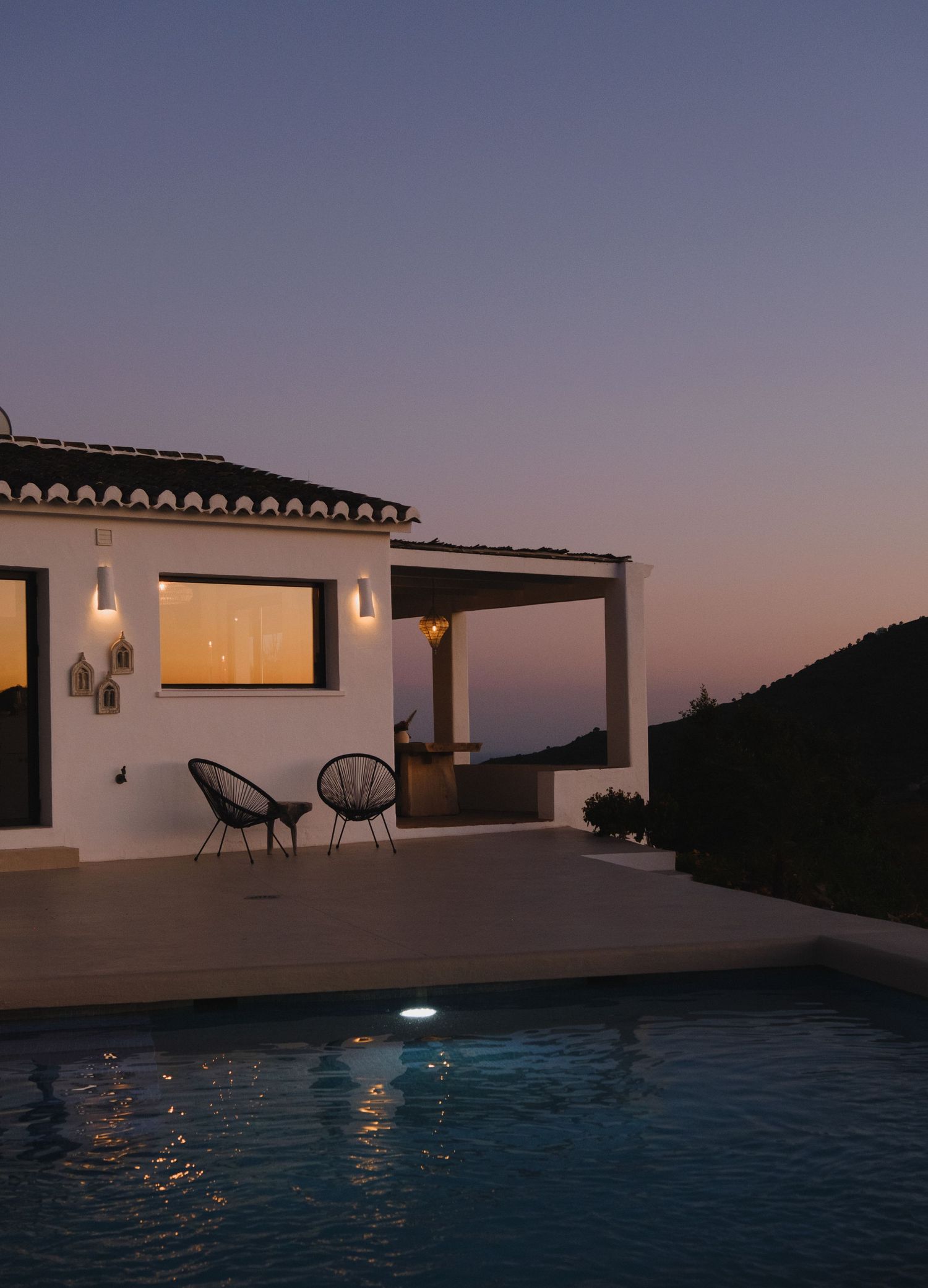 White Mediterranean villa with illuminated pool at dusk, featuring outdoor seating and pergola against a twilight sky.