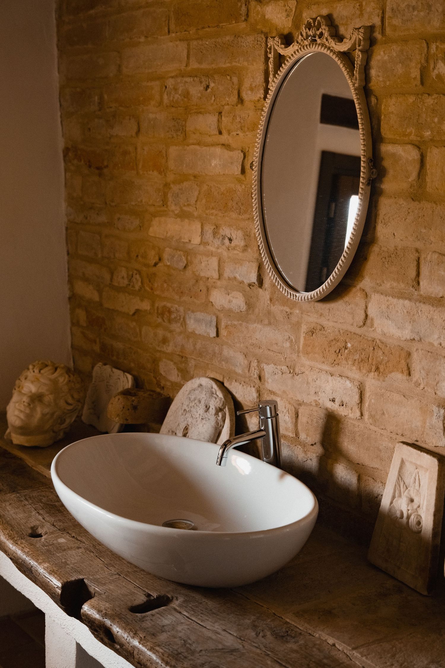 Rustic bathroom with oval vintage mirror, white vessel sink on wooden vanity, and exposed brick wall.