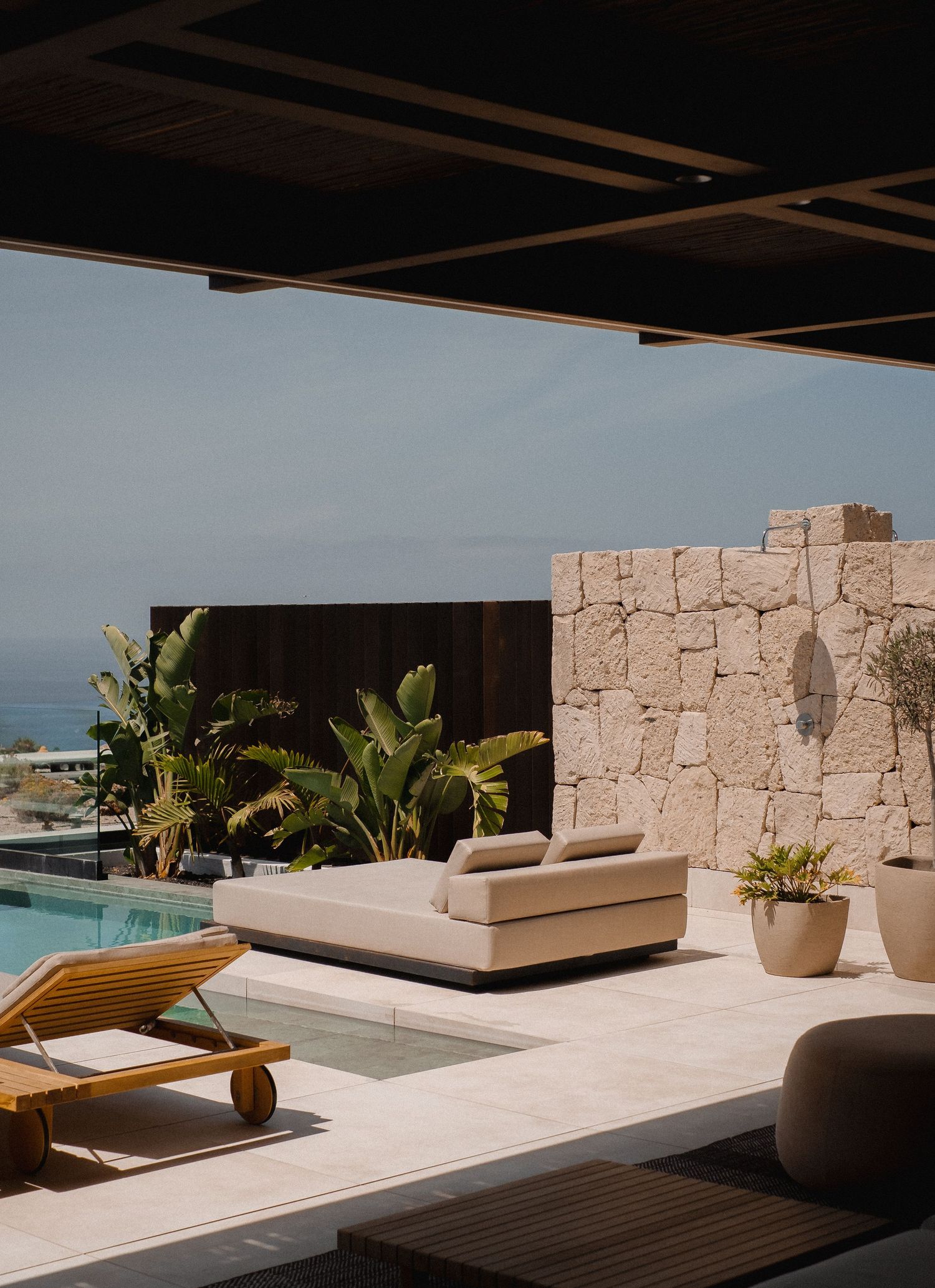 Luxury outdoor terrace with pool, stone wall, tropical plants, and modern lounge furniture overlooking the ocean.