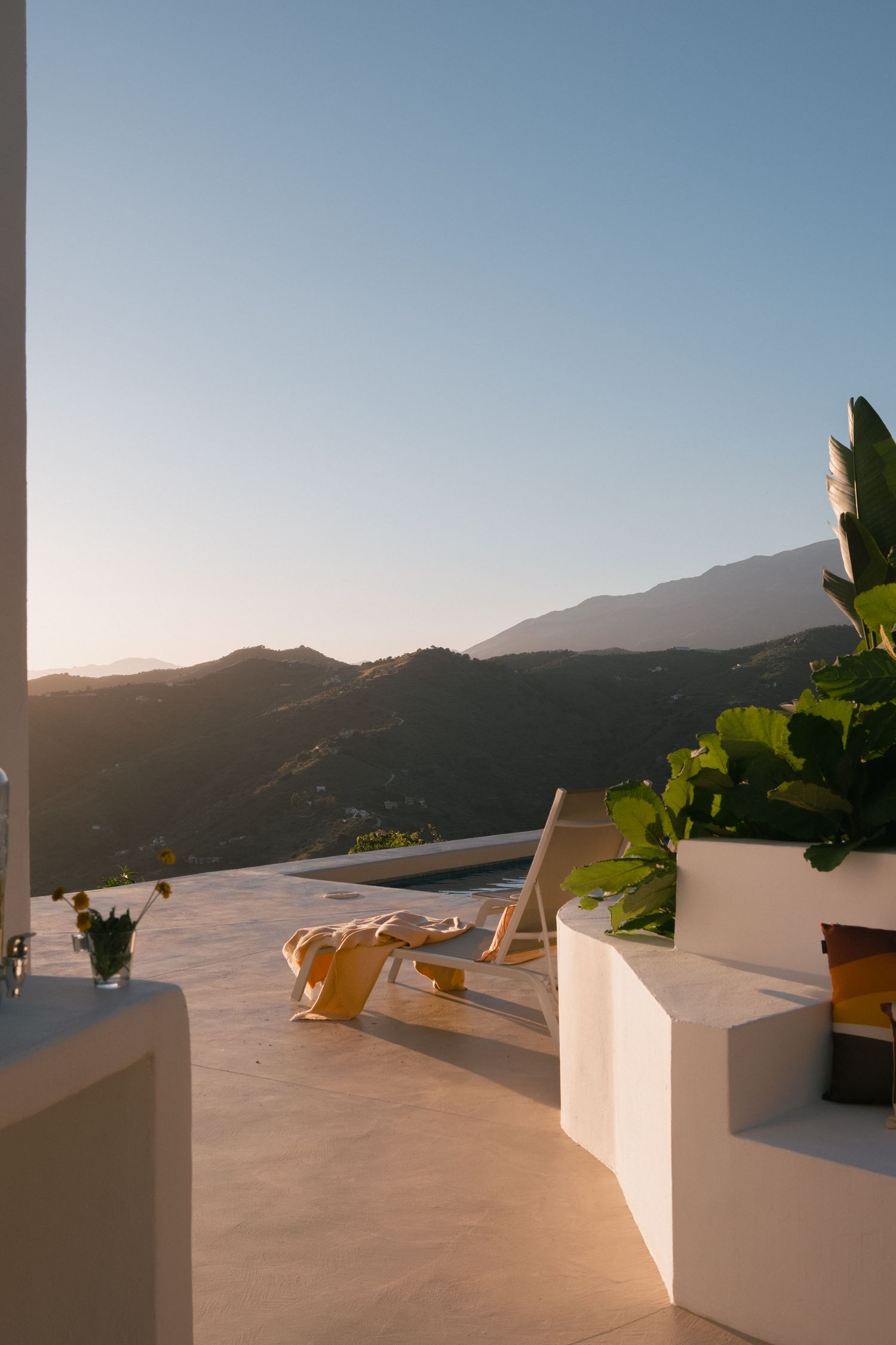 Luxury terrace with lounge chair overlooking mountain sunset, white architecture, tropical plants, clear blue sky.