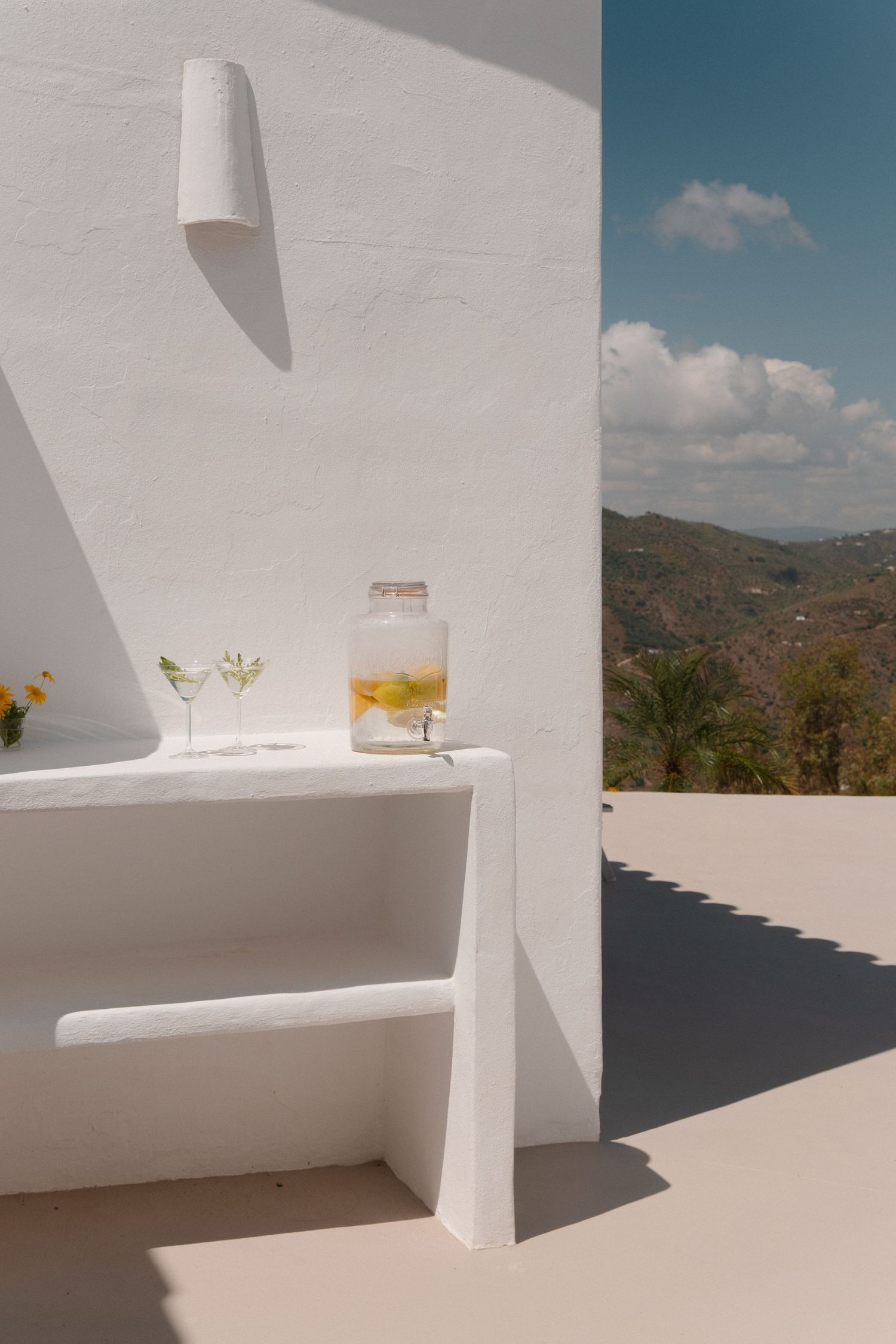 White Mediterranean terrace with lemonade pitcher and cocktail glasses, overlooking scenic mountain landscape under blue sky.