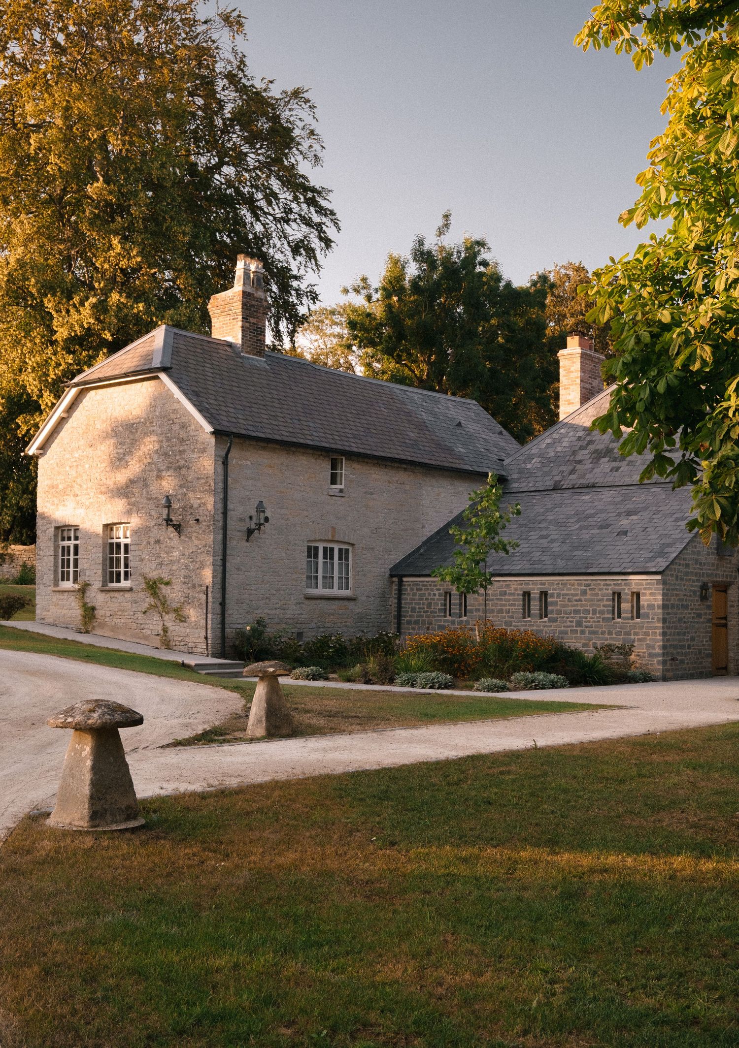 A charming stone Cotswold cottage with slate roof, chimneys, and lush garden surroundings bathed in warm golden evening light.