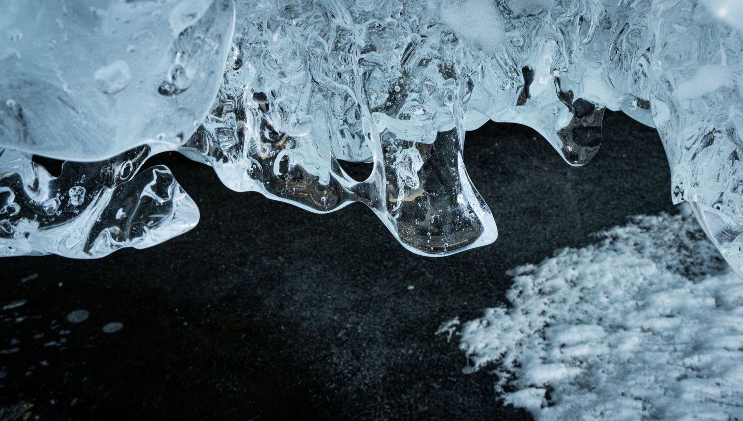 Close-up of clear ice formations resting on black volcanic sand along an icy shoreline.