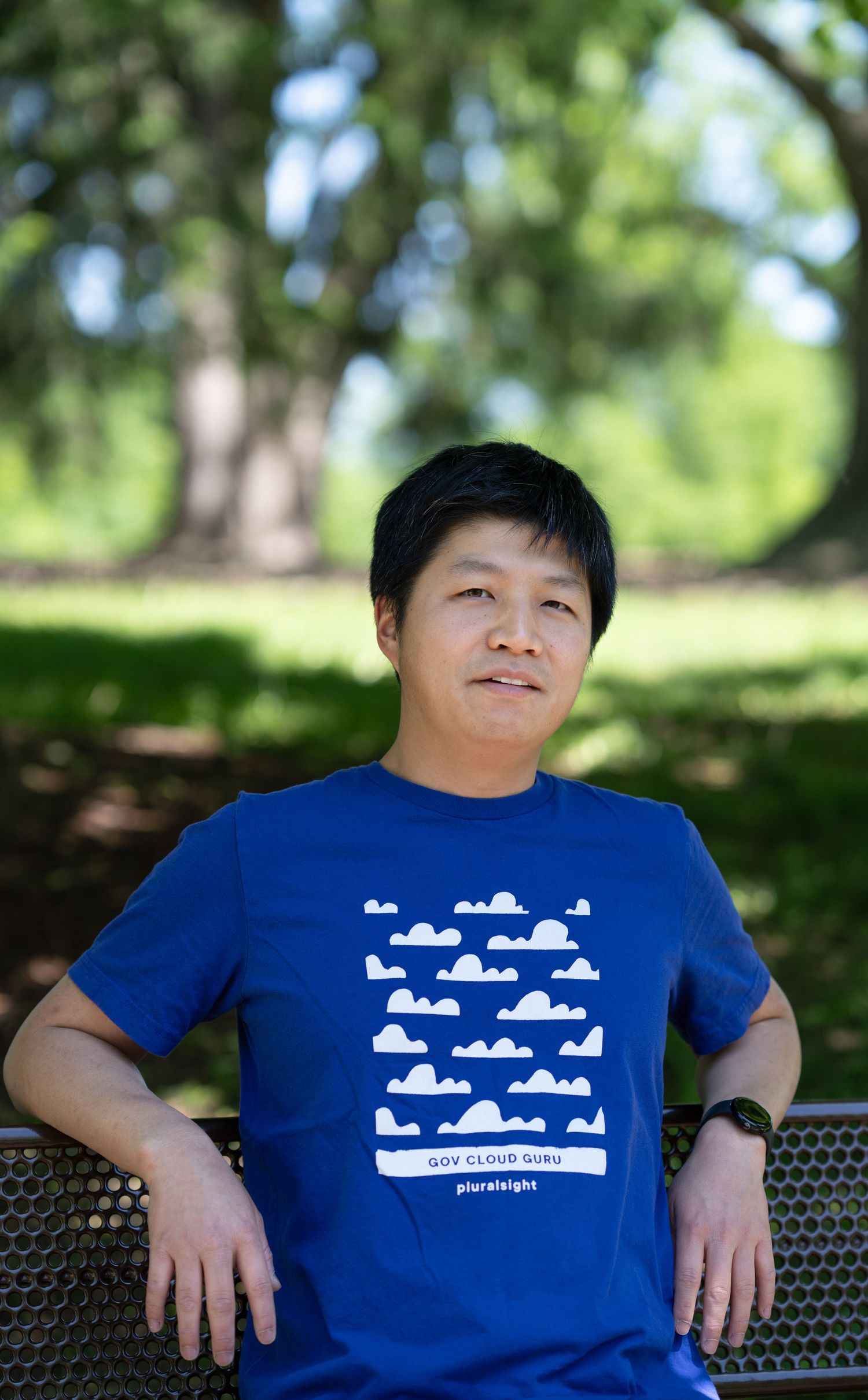 Casual outdoor portrait of a man seated on a park bench surrounded by trees and natural light.