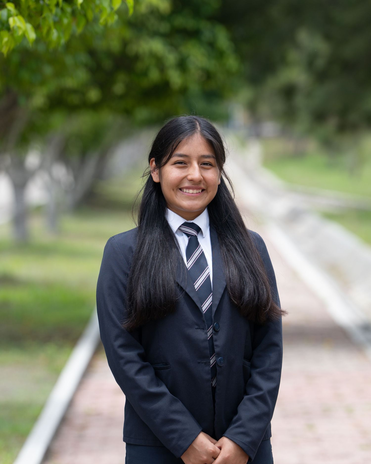 Student portrait outdoors wearing a formal blazer and tie, smiling along a tree-lined pathway.