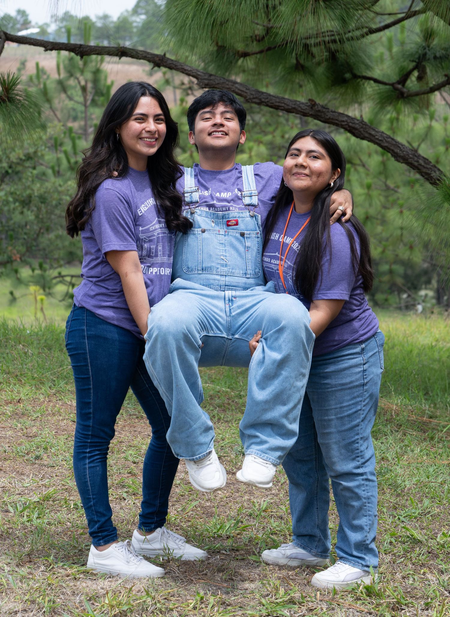 Playful outdoor group portrait of three friends laughing while lifting one person in a wooded setting.