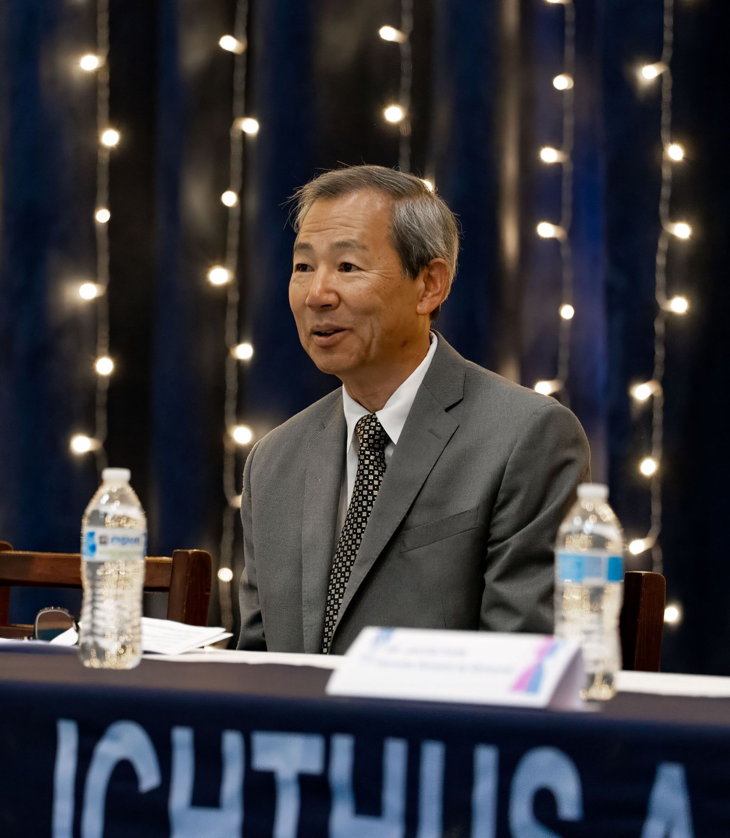 Speaker seated at a table during a formal event, smiling while speaking in front of soft string lights.