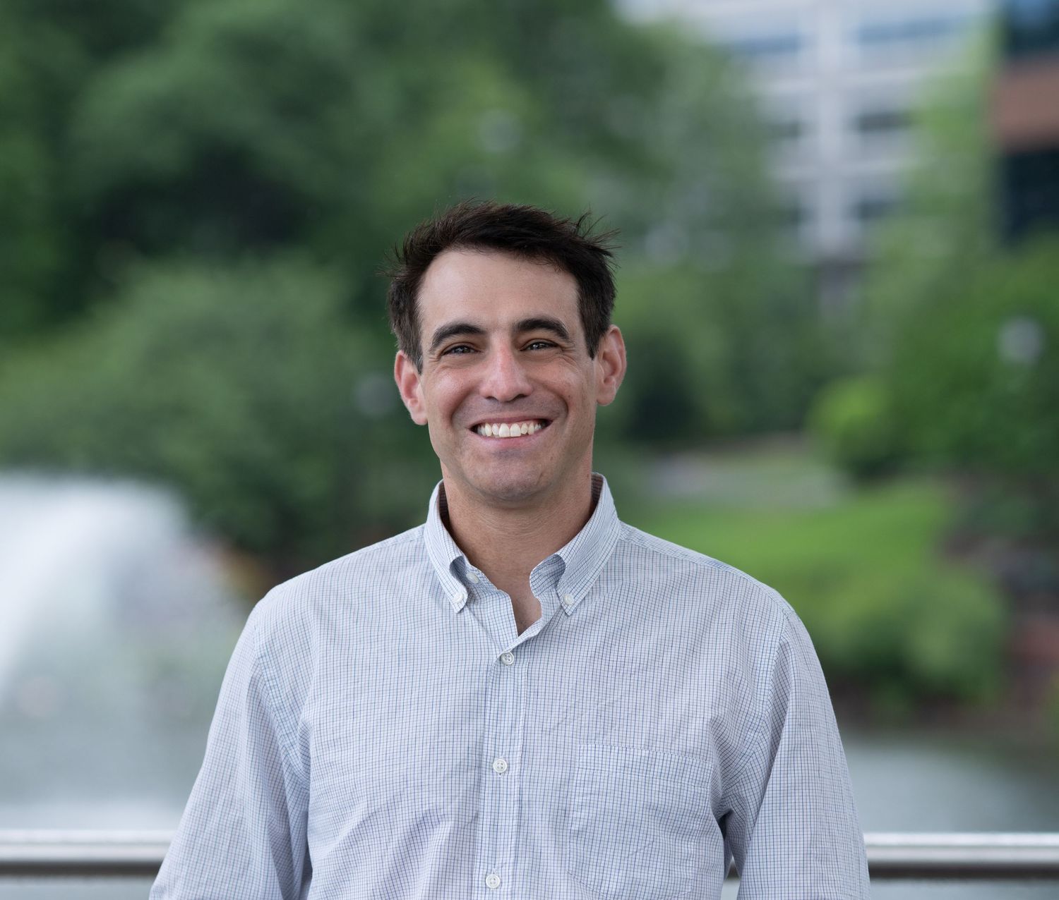 Outdoor professional headshot of a smiling man standing near a fountain with greenery and city buildings softly blurred in the background.