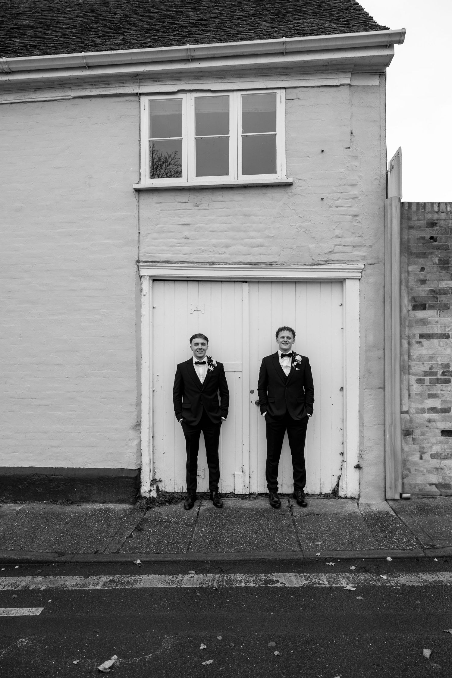 Two people in formal black suits stand symmetrically against white double doors in a black and white photo.