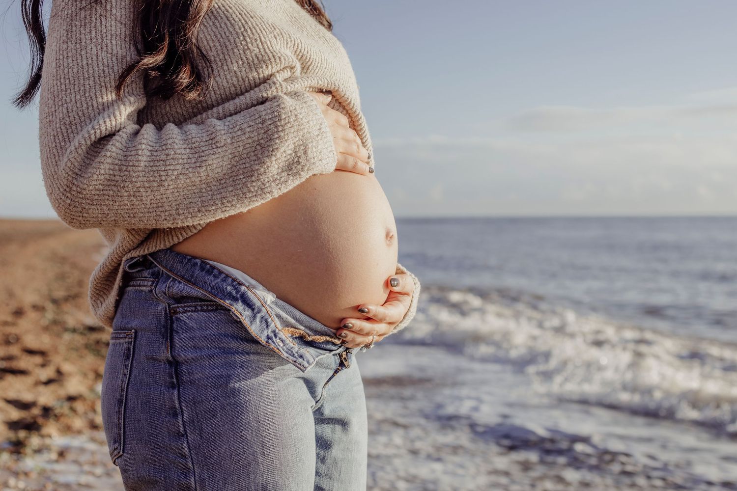 Person wearing knit sweater showing pregnant belly against ocean backdrop.
