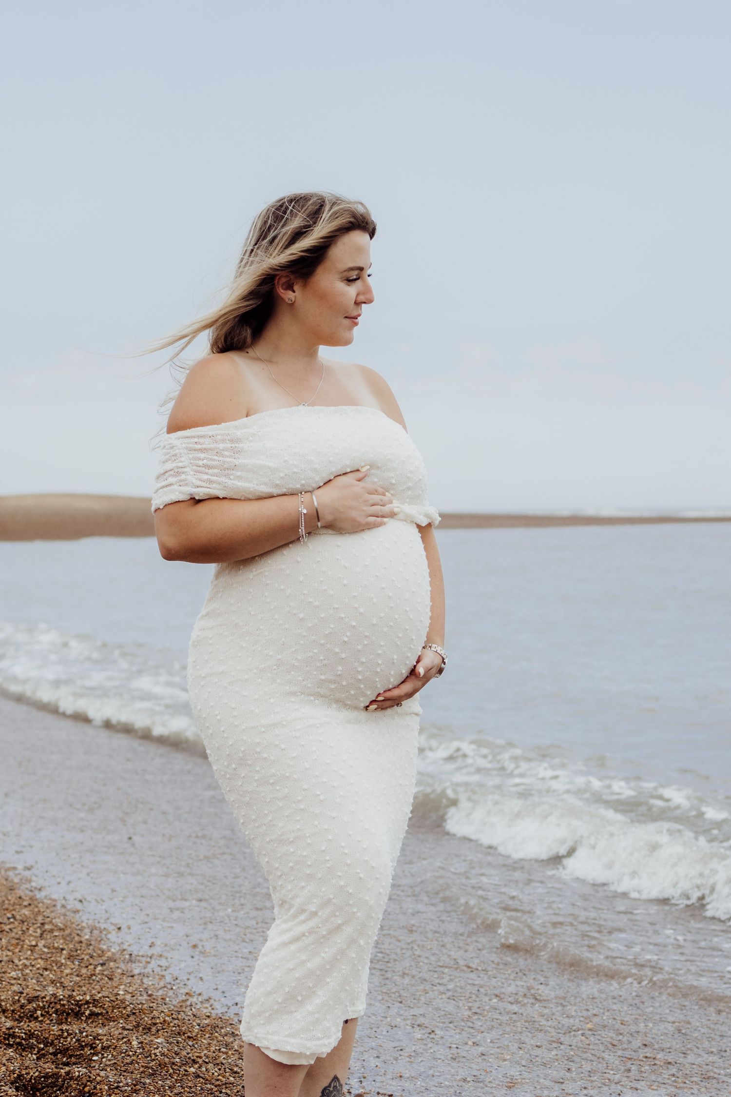 A pregnant woman in a white lace dress poses thoughtfully by the ocean shore as gentle waves roll in.