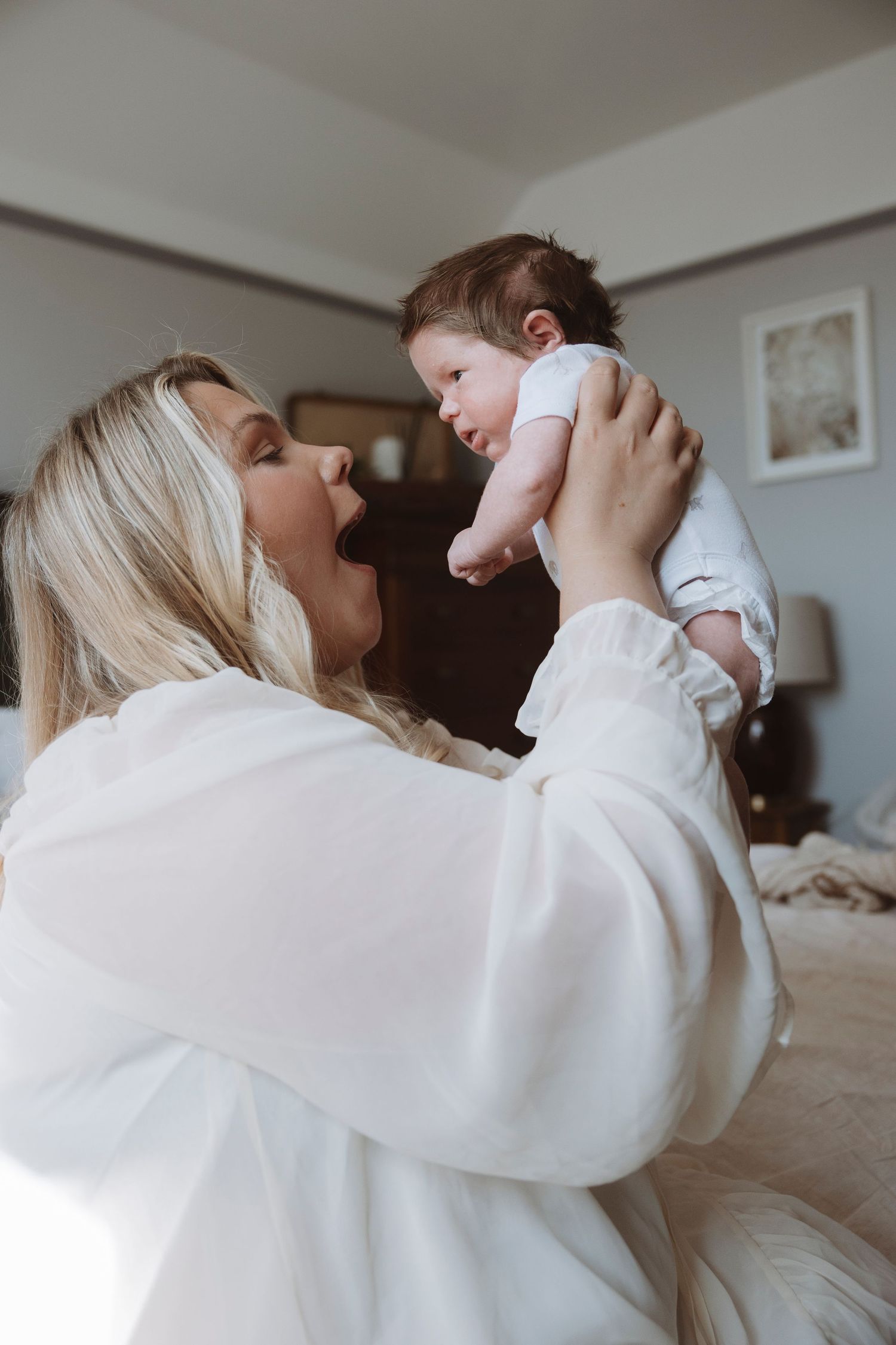 A joyful blonde woman in a white robe lifts her newborn baby above her head while sitting on a bed.