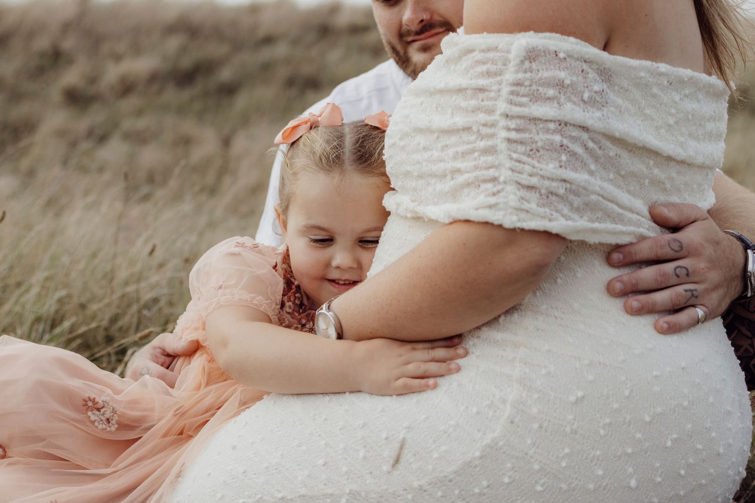 A tender maternity photo session in a grassy field at sunset with soft lighting and neutral tones.