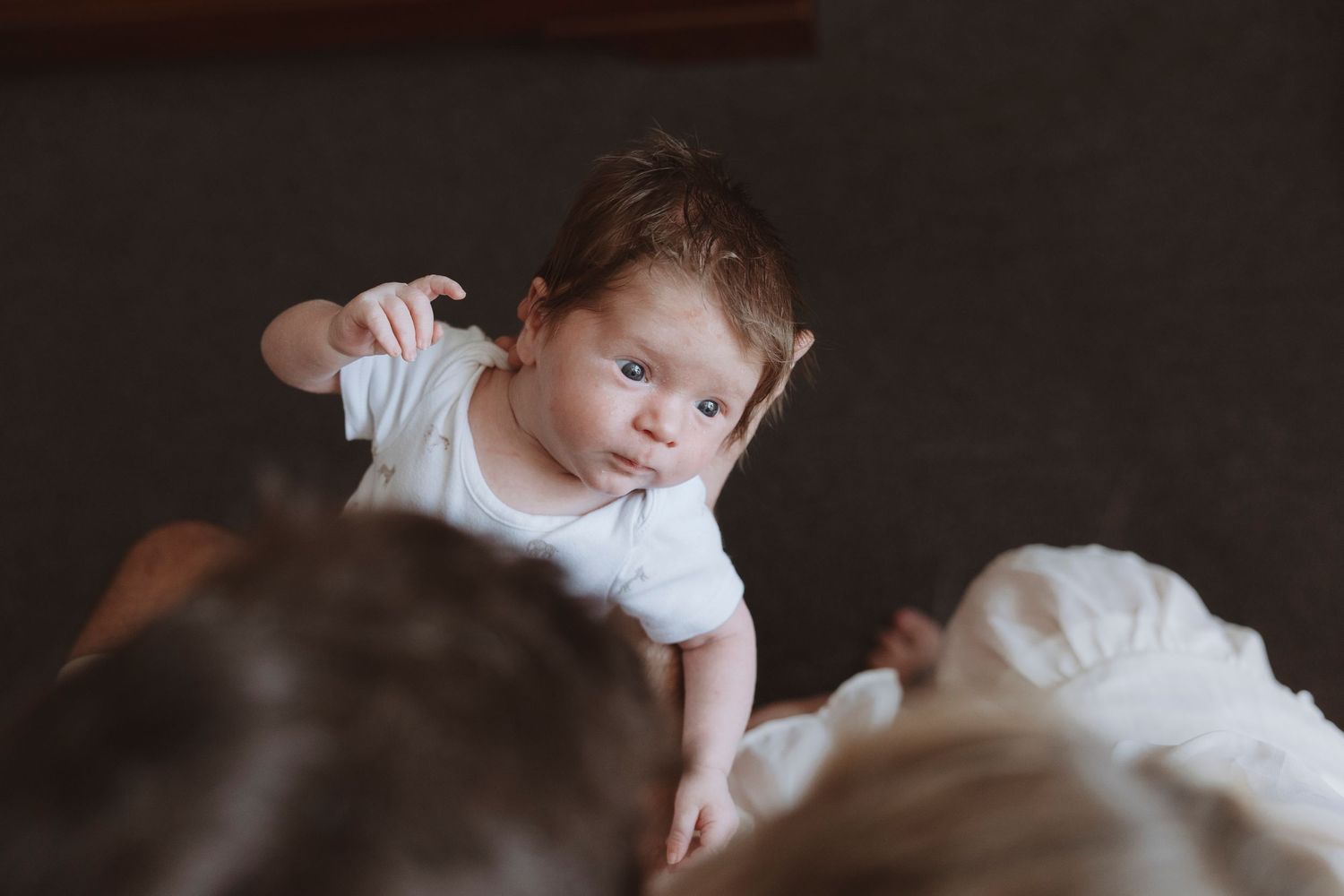 A curious newborn baby in a white onesie reaches out while being held by parents in a softly lit room.