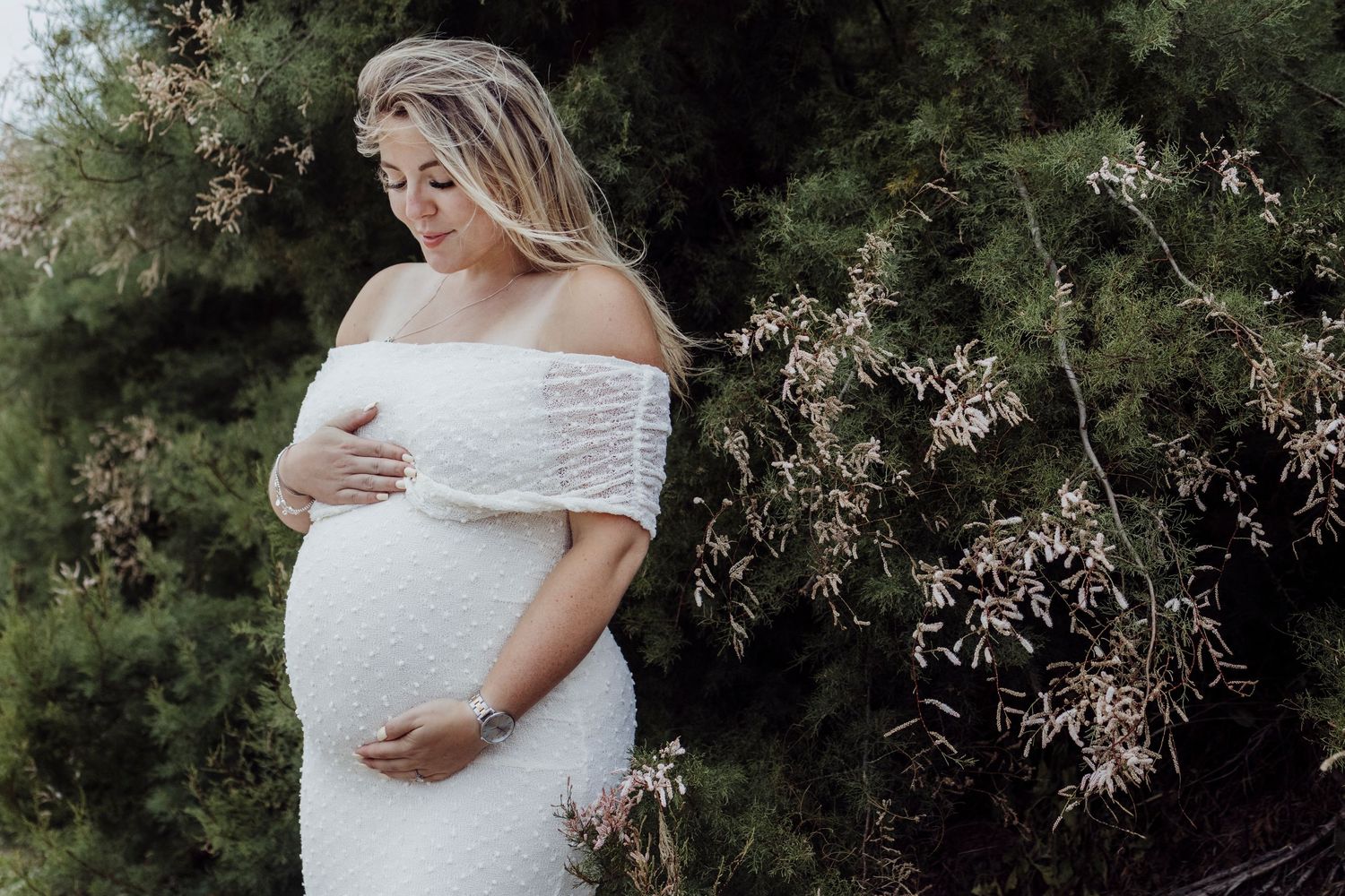 Expectant mother in white lace off-shoulder dress poses outdoors against greenery in soft natural light.