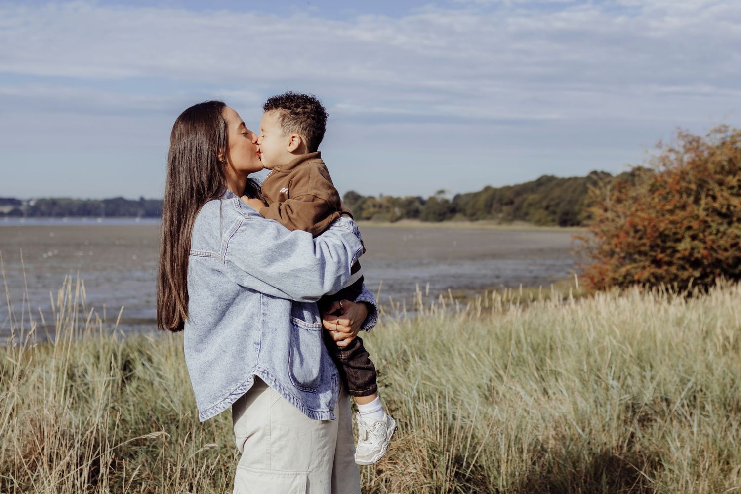 Mother and son share a tender moment in a coastal meadow with tall grass and blue sky.