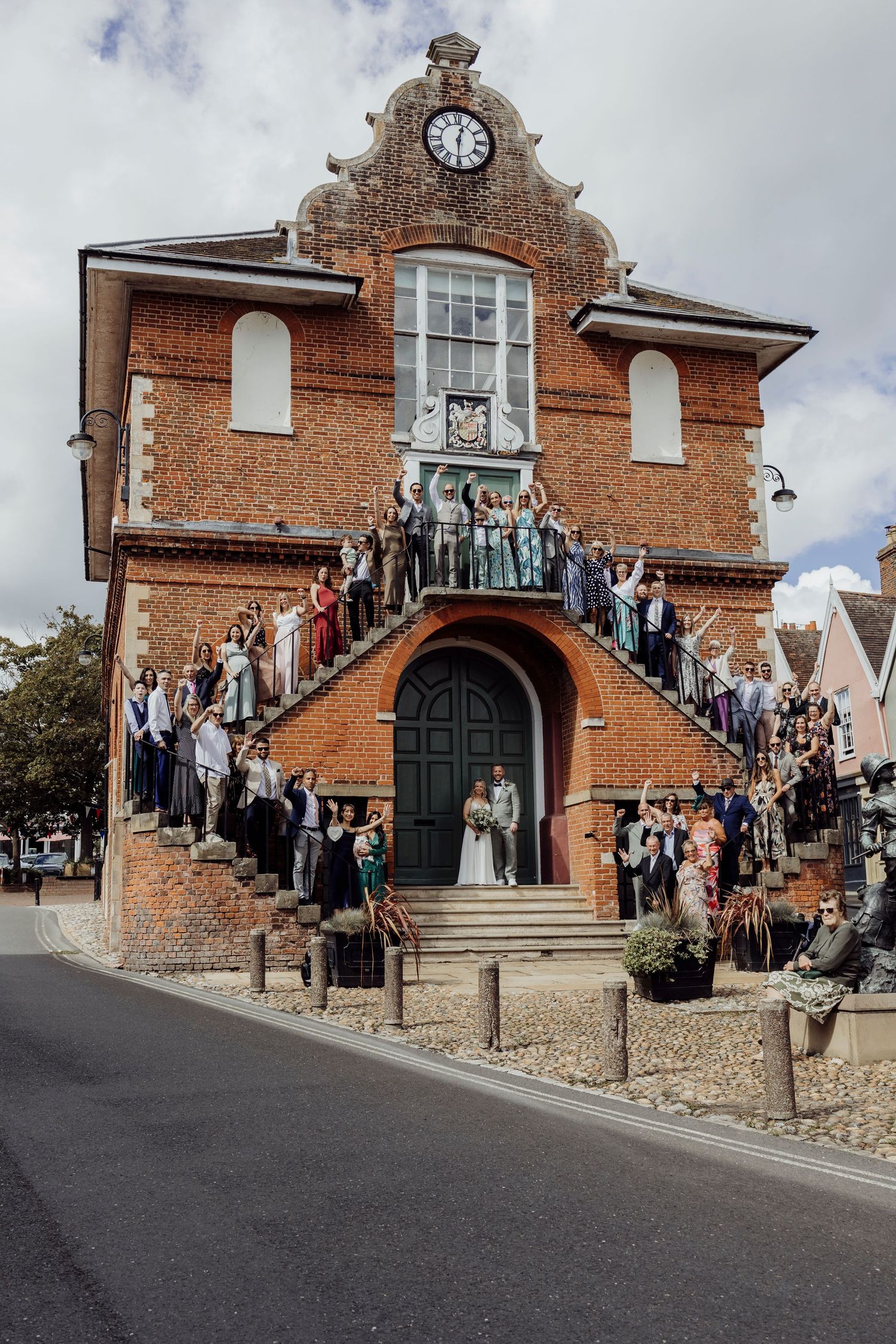 Large wedding party poses on the steps of a historic brick clock tower building.