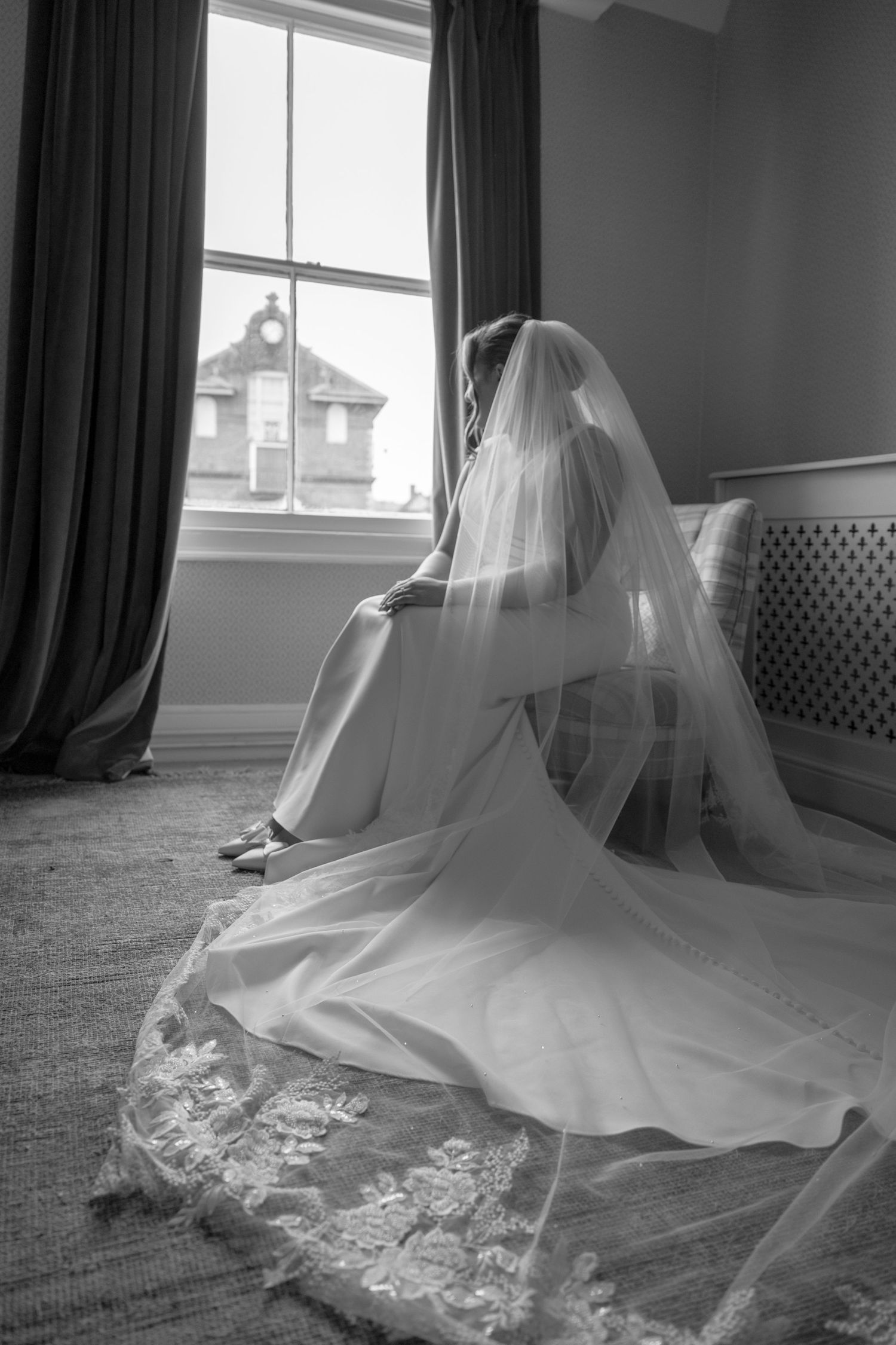 A bride in flowing veil sits by a window in dramatic black and white lighting.
