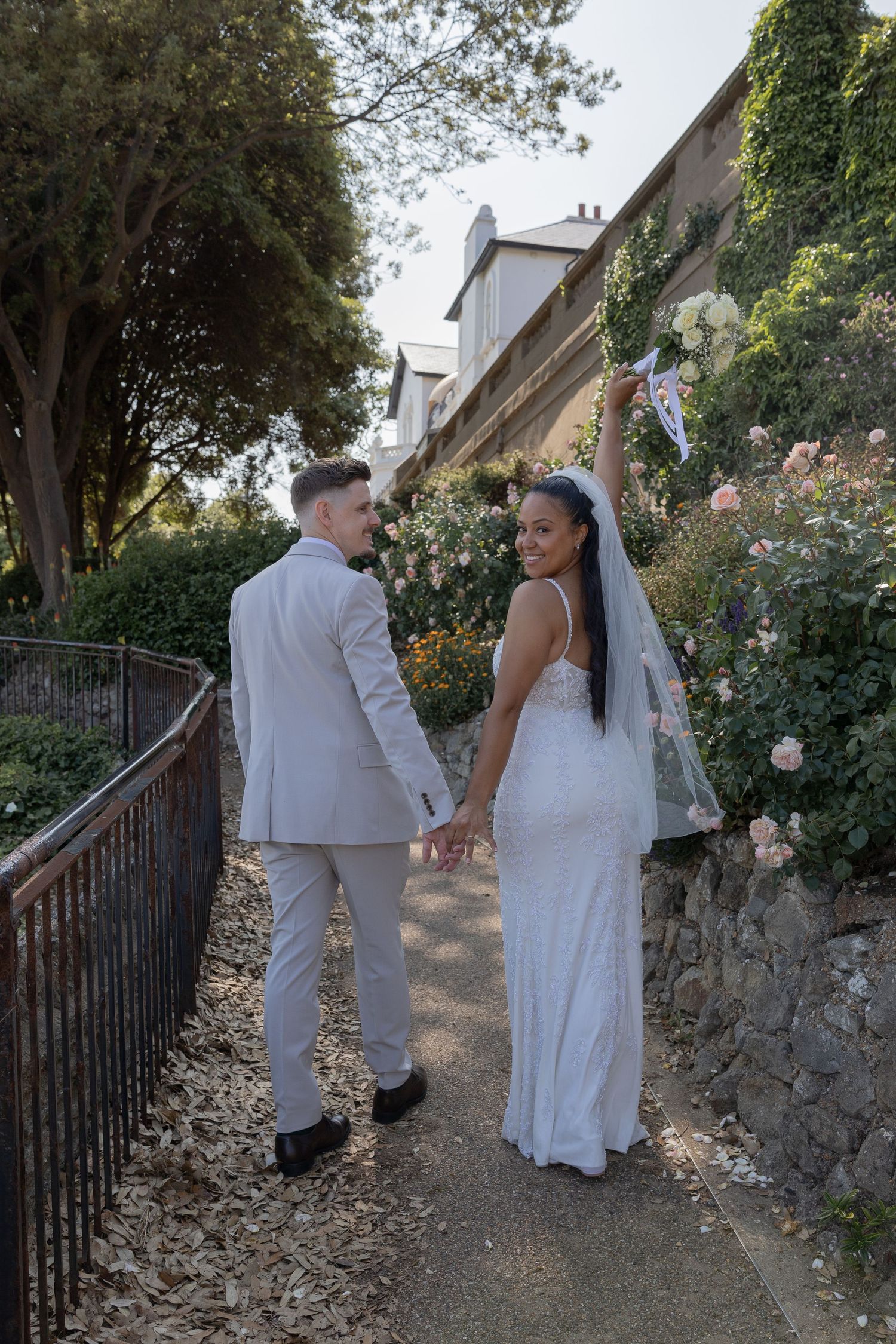 A couple in white wedding attire walks along a garden path with stone walls and greenery.