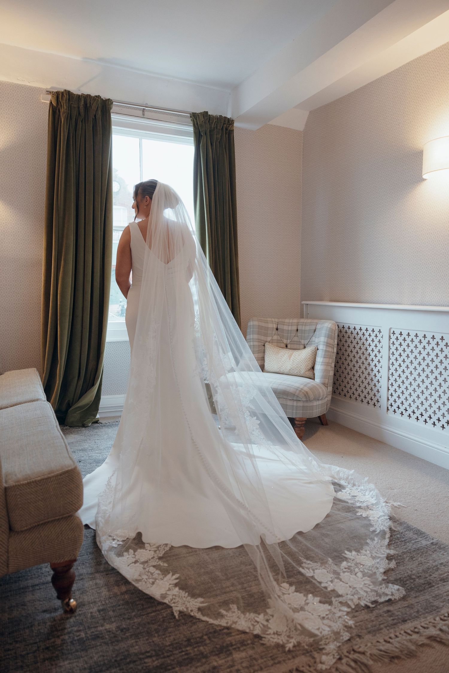 A bride in a flowing lace veil and white dress stands near elegant curtains in a bridal suite.