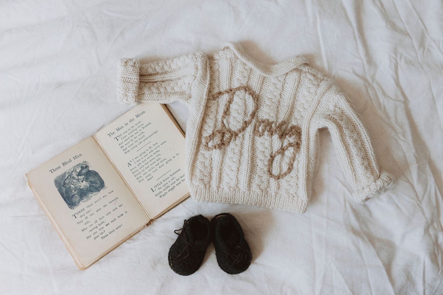 Cream knit baby sweater with name embroidery, vintage book, and brown baby shoes on white bedding.