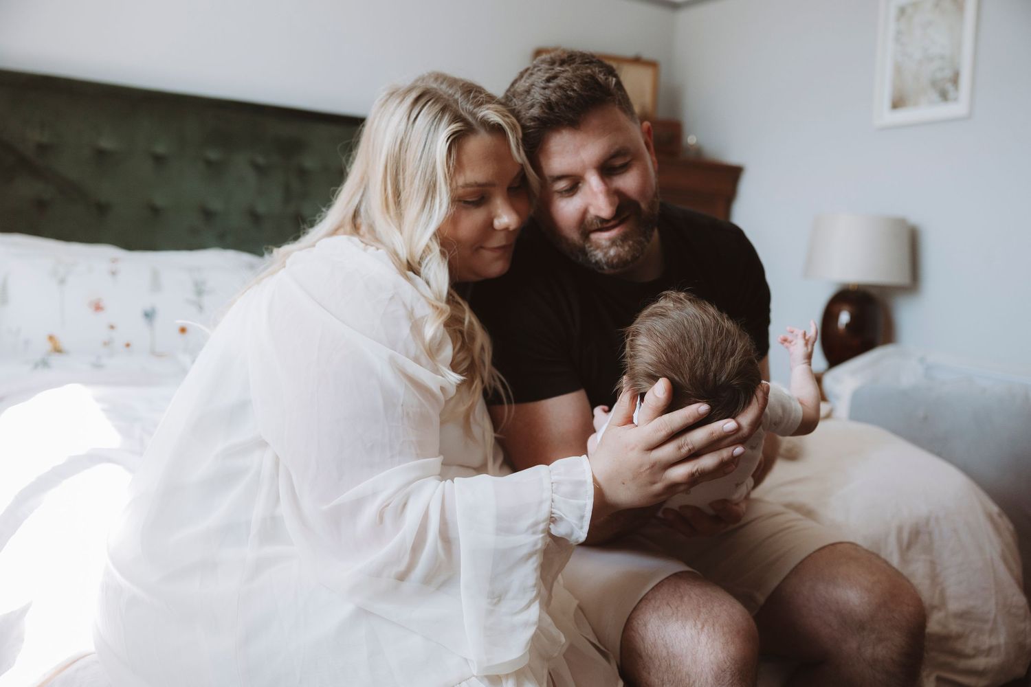 A couple sits on a bed, lovingly cradling their newborn baby in a bright, cozy bedroom.