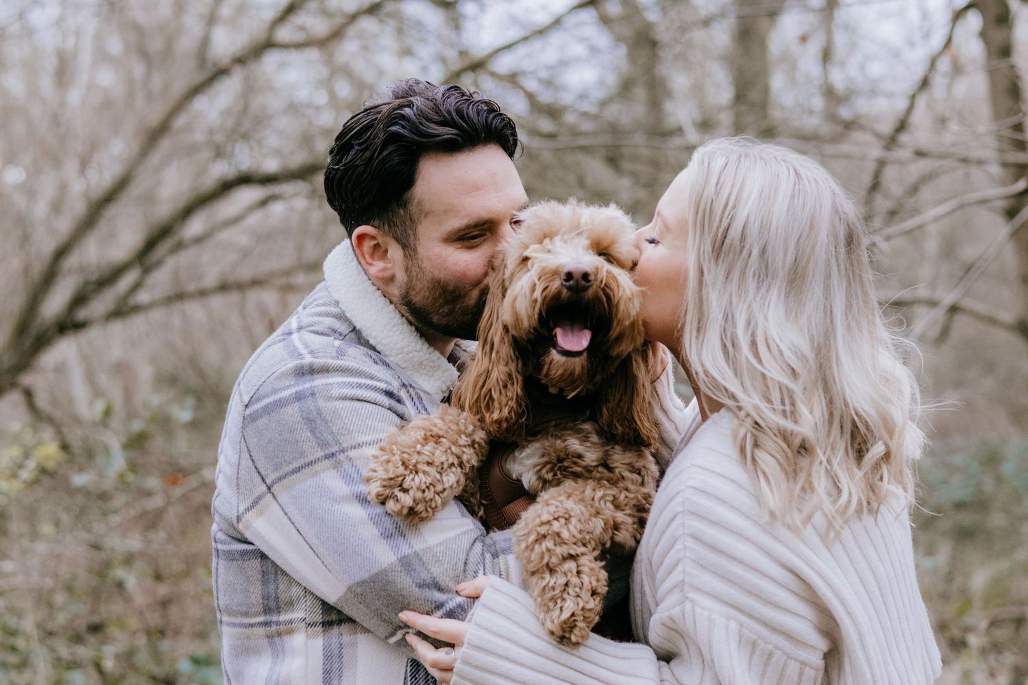 A couple and their goldendoodle dog share tender moments in a winter forest setting.