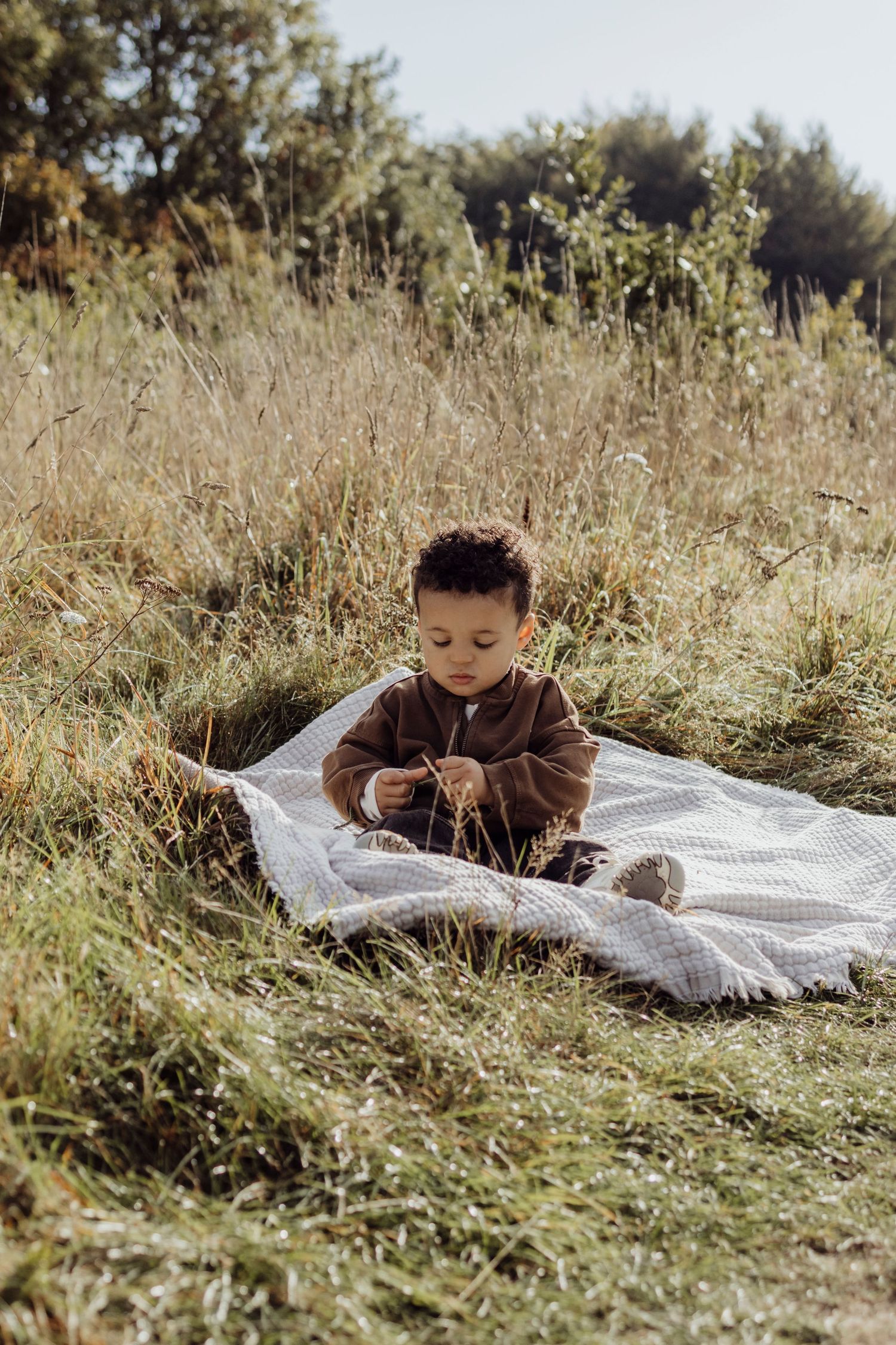 A small child lies on a white blanket surrounded by tall grass in a natural outdoor setting.