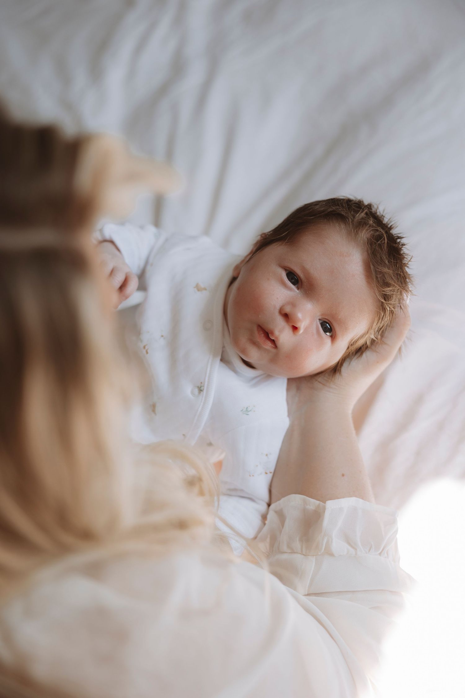 A newborn baby in white onesie lying on white bedding, gazing up at mother in soft natural light.