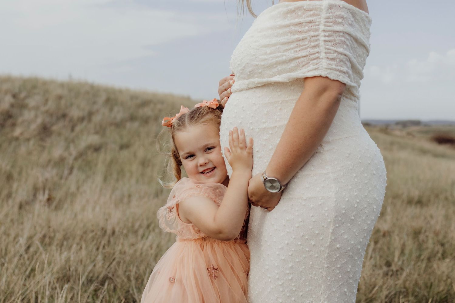 A mother in a white dress holds her child wearing a peach dress in a grassy field during sunset.