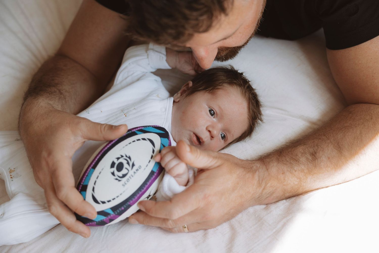 Father kissing newborn baby holding a rugby ball while lying on a white bed.