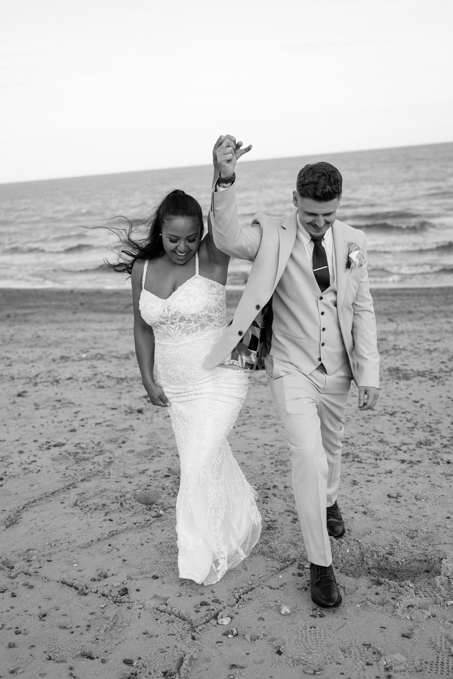 A couple strolls along a sandy beach shore in elegant wedding attire.