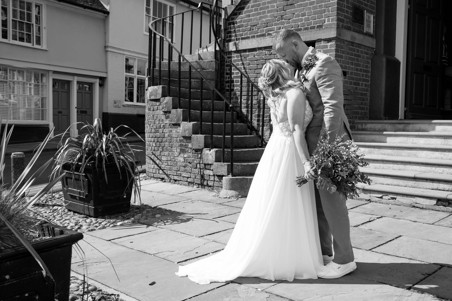 A couple in wedding attire poses on a historic brick-lined street with classic architecture.