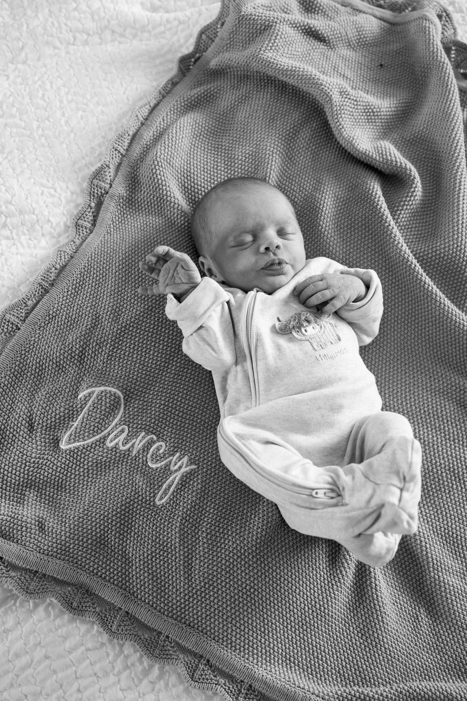 A sleeping newborn baby swaddled in white clothing rests on a textured gray blanket in a black and white photograph.