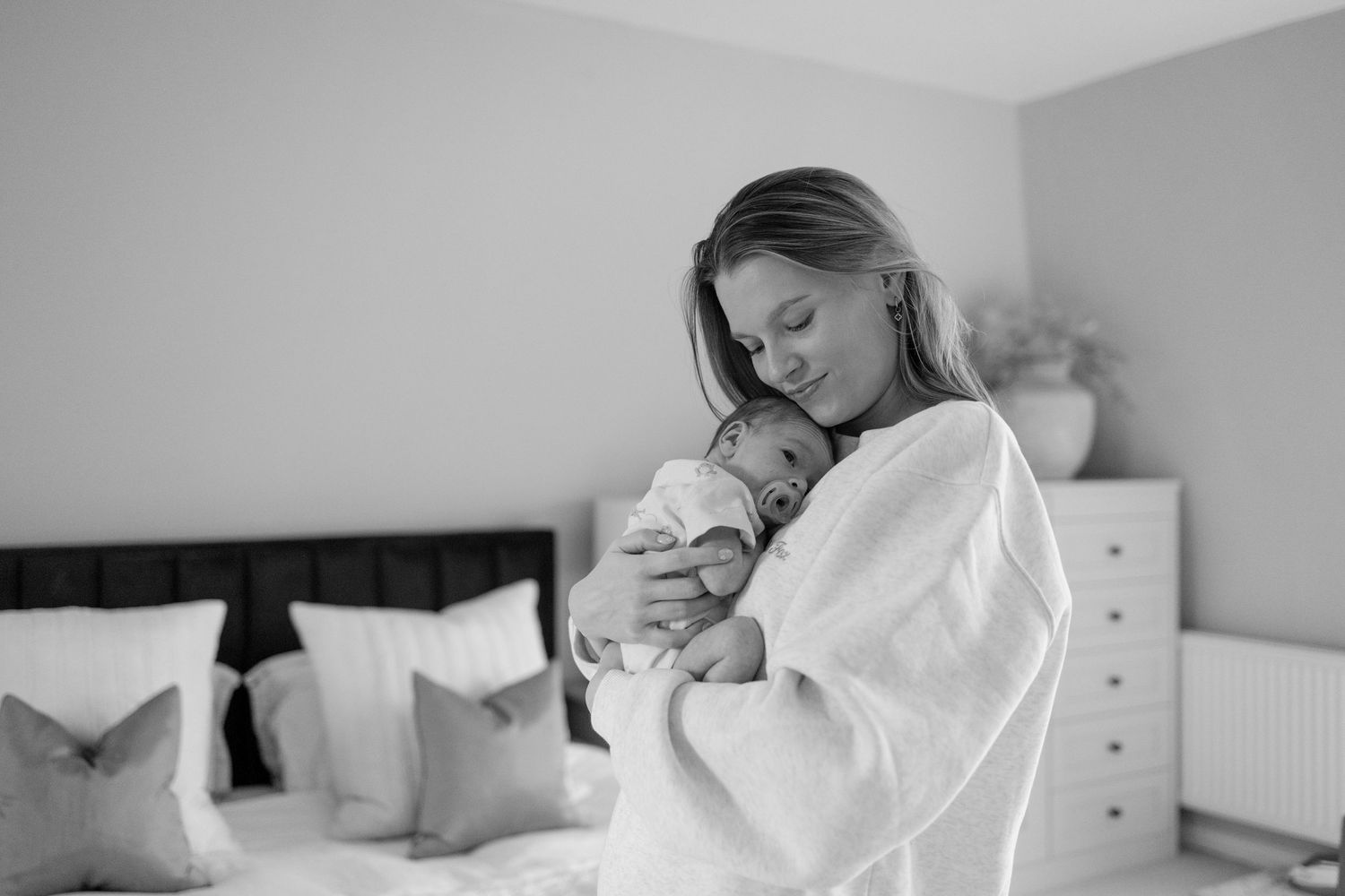 A tender black and white moment in a bedroom as a parent cradles a newborn baby while wearing a cozy white robe.