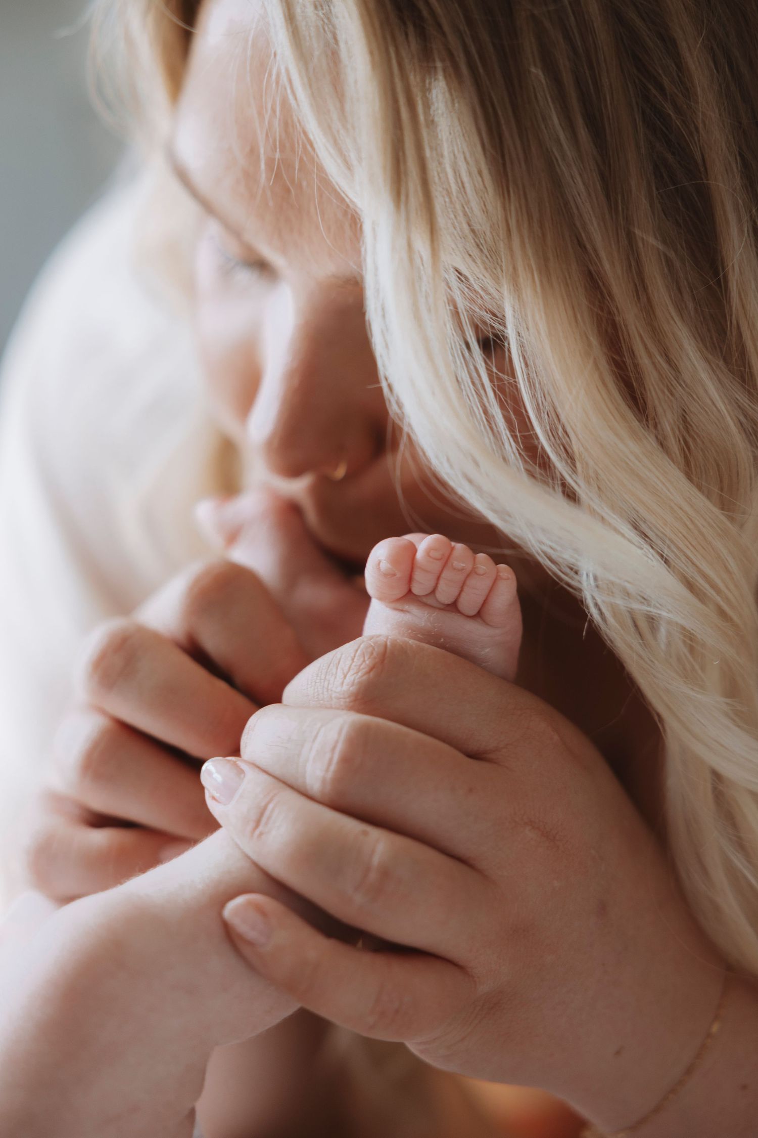 A blonde mother tenderly holds her newborn baby's tiny feet close to her face in a warm, intimate moment.