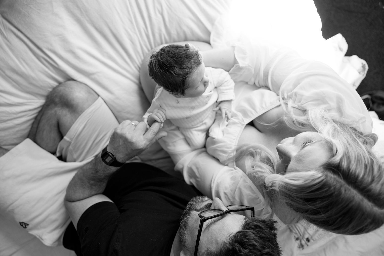 Black and white photo of parents lying on bed with newborn baby, sharing a tender family moment.