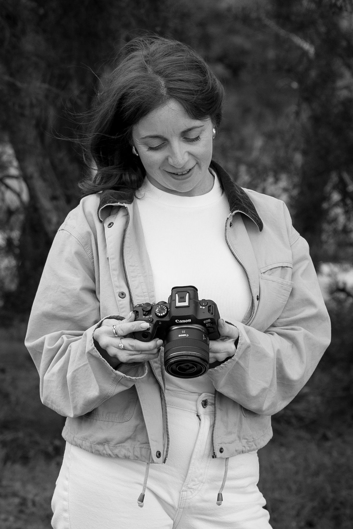 Young woman smiling while reviewing photos on a Canon camera outdoors, wearing a beige jacket, black and white photo.