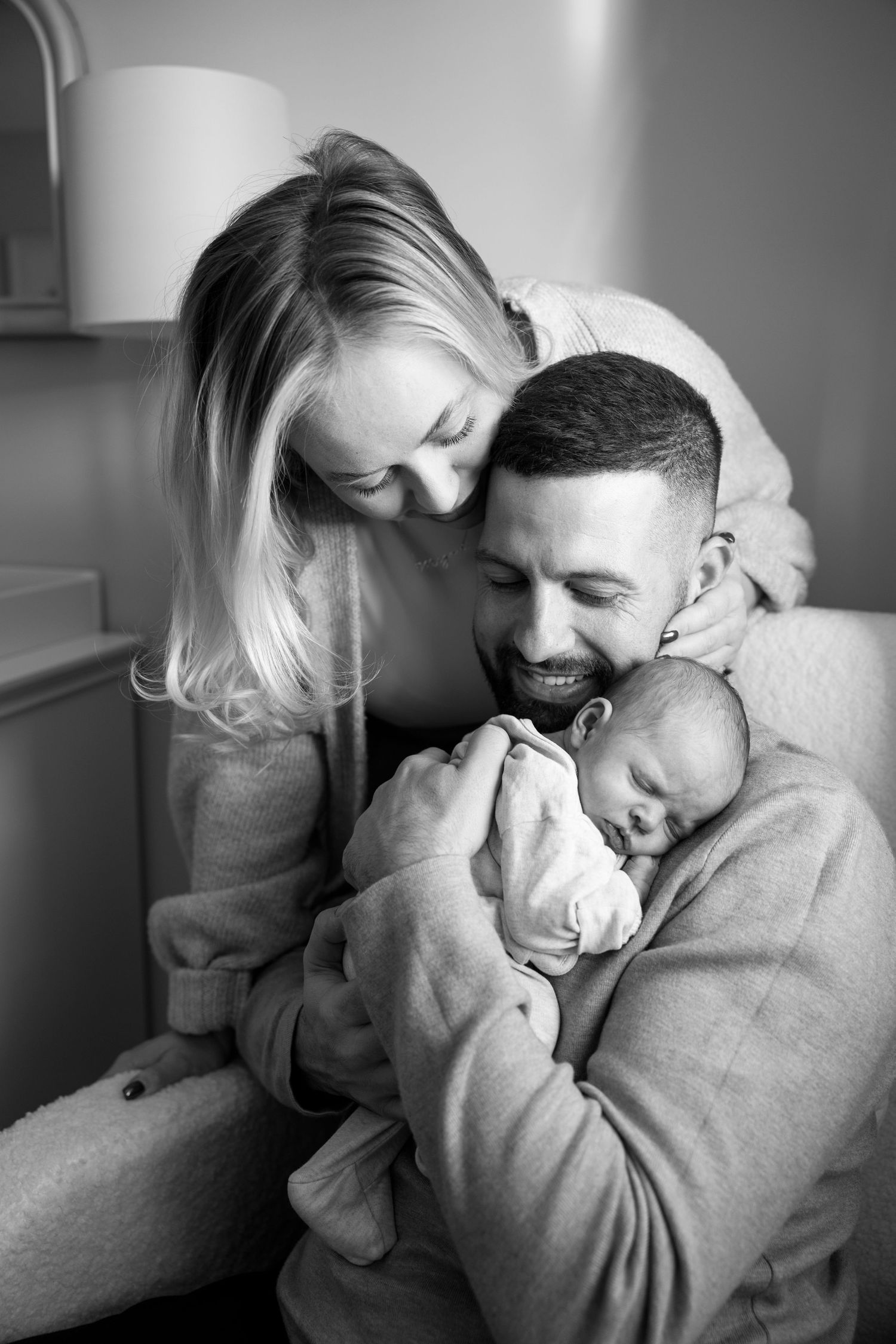 A tender black and white family moment as parents lovingly embrace their sleeping newborn baby.