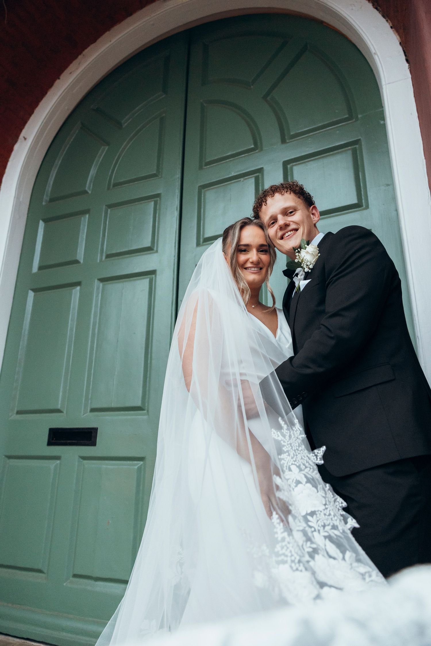 A couple poses together in front of a large mint green door on their wedding day.