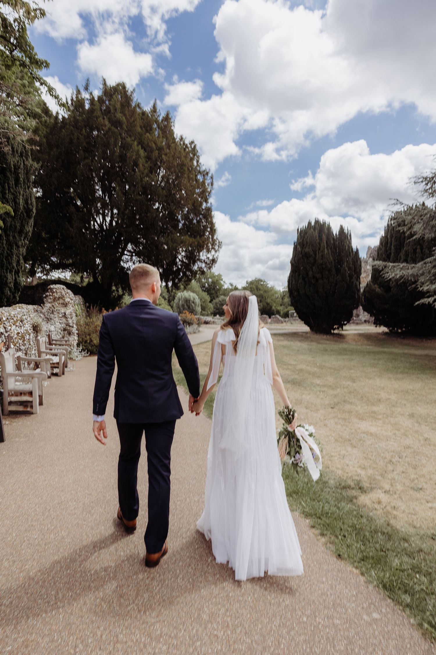 A bride and groom walk hand in hand down a garden path on a sunny day.