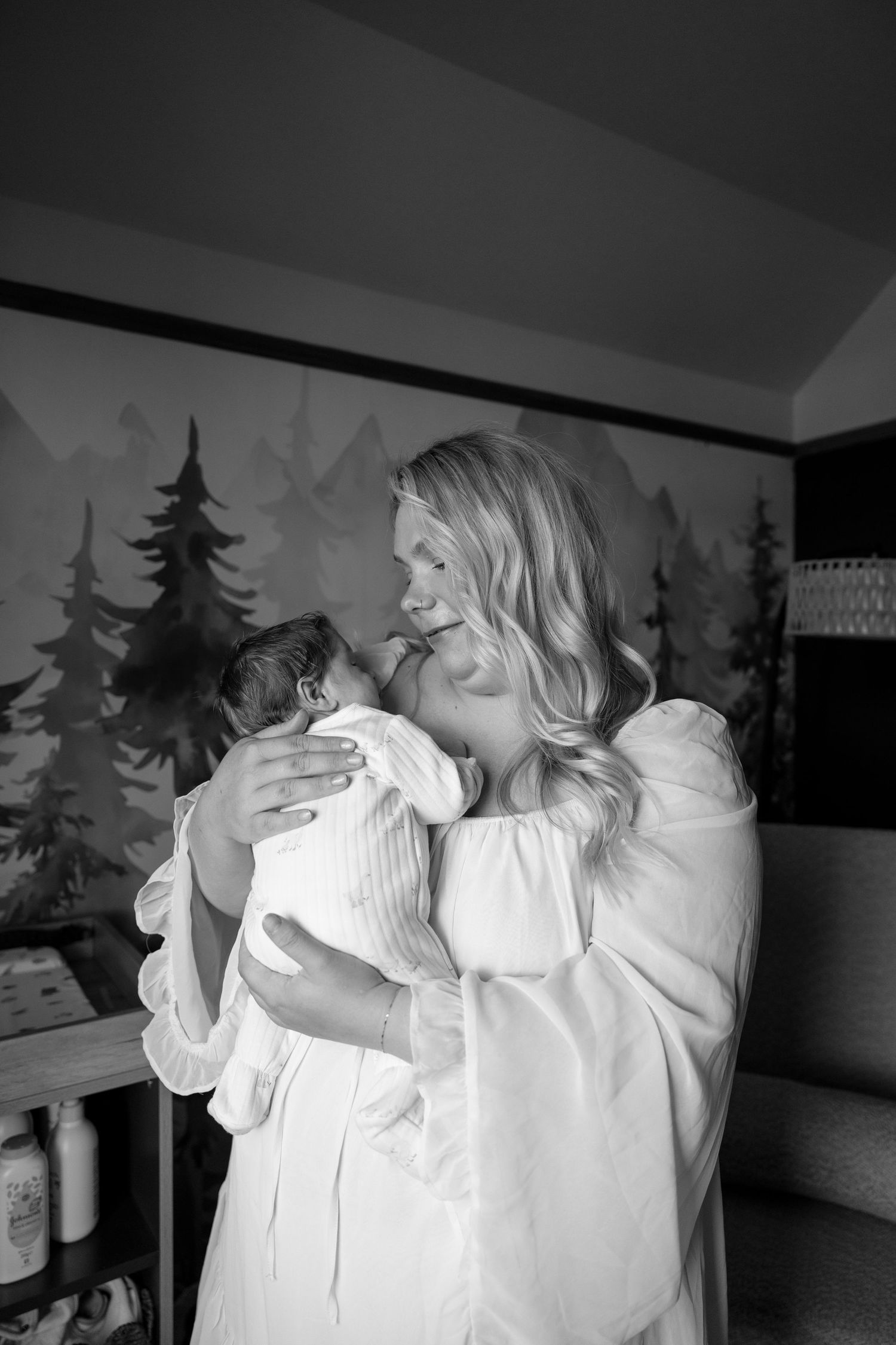 Black and white photo of a smiling blonde woman in a white dress tenderly holding a newborn baby in a nursery.