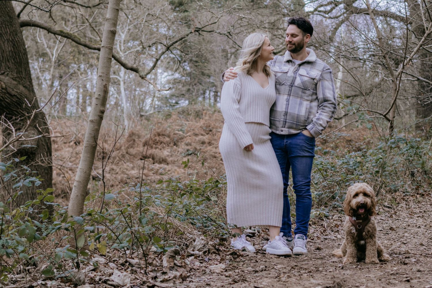 A couple and their dog walk through a serene wooded area during an engagement photoshoot in autumn.