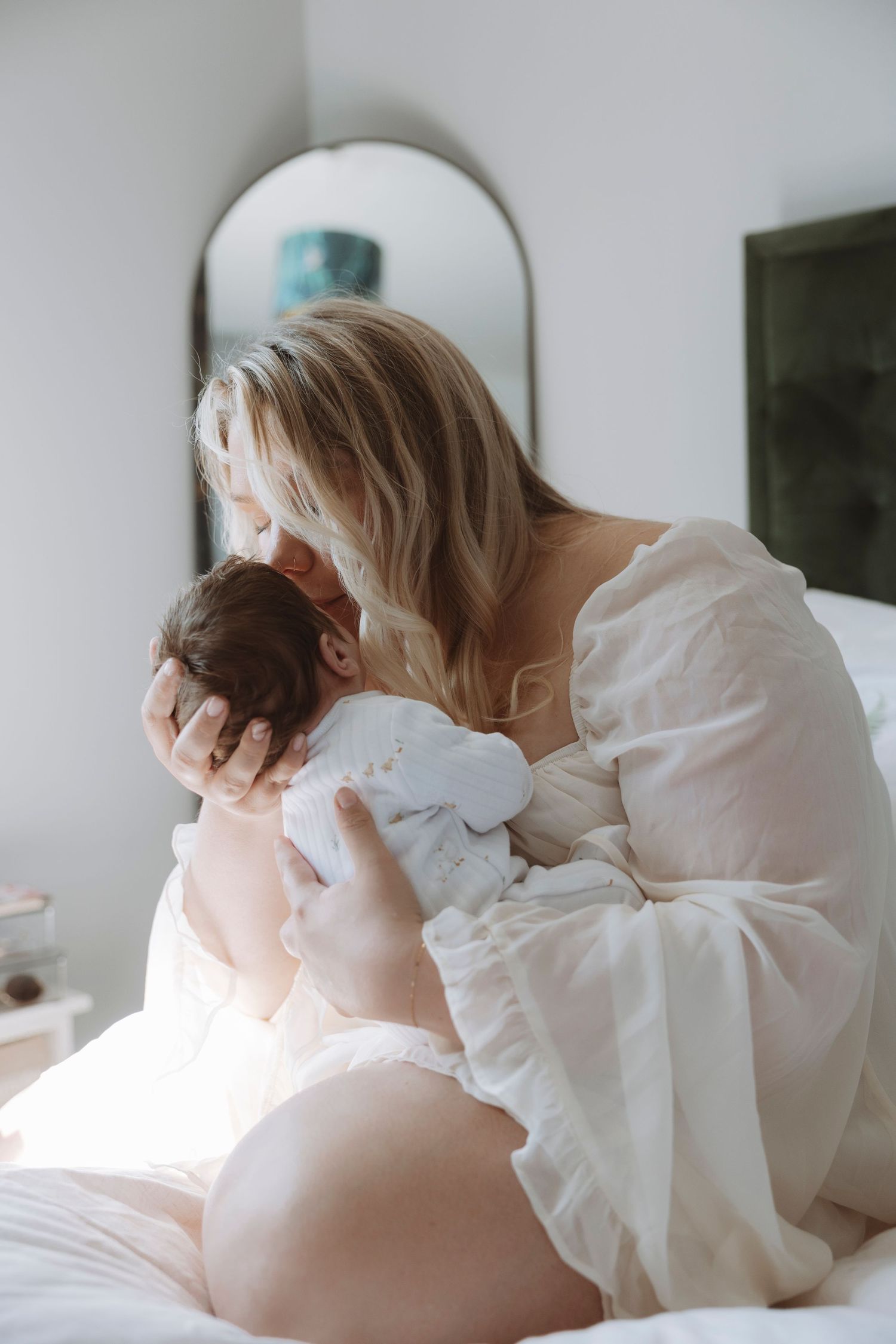 A blonde mother in a white dress tenderly holds and kisses her newborn baby while sitting on a bed.