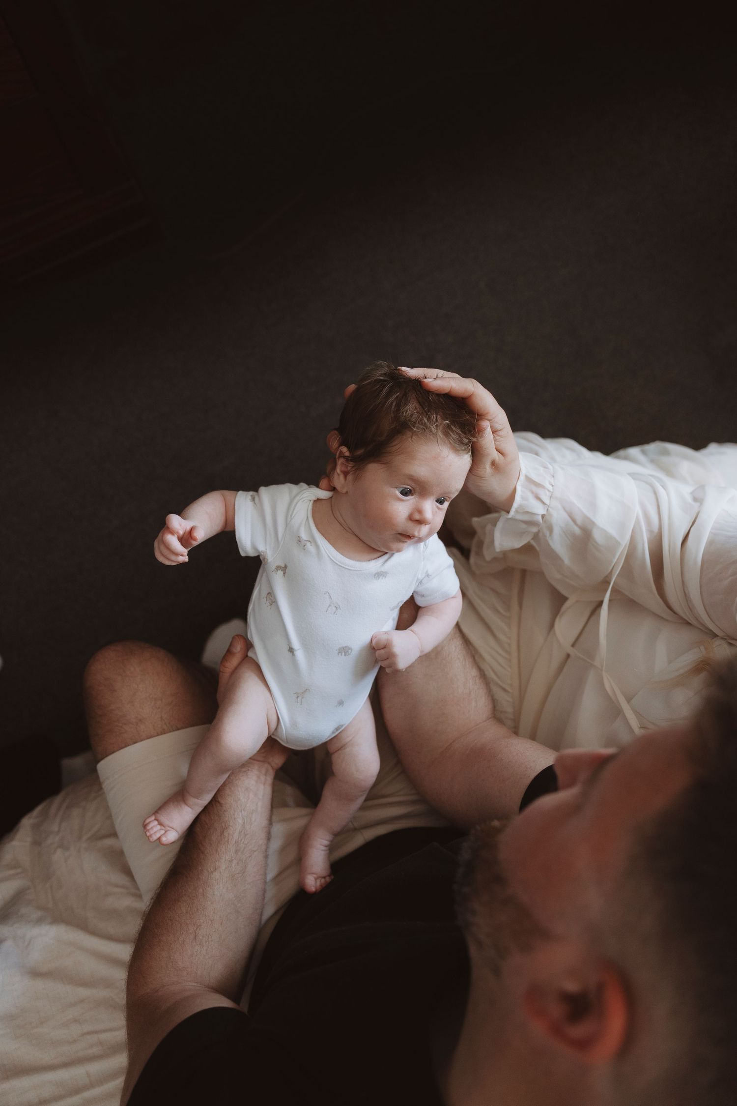 Father gently holds newborn baby in white onesie on bed, tenderly touching infant's head in cozy home setting.