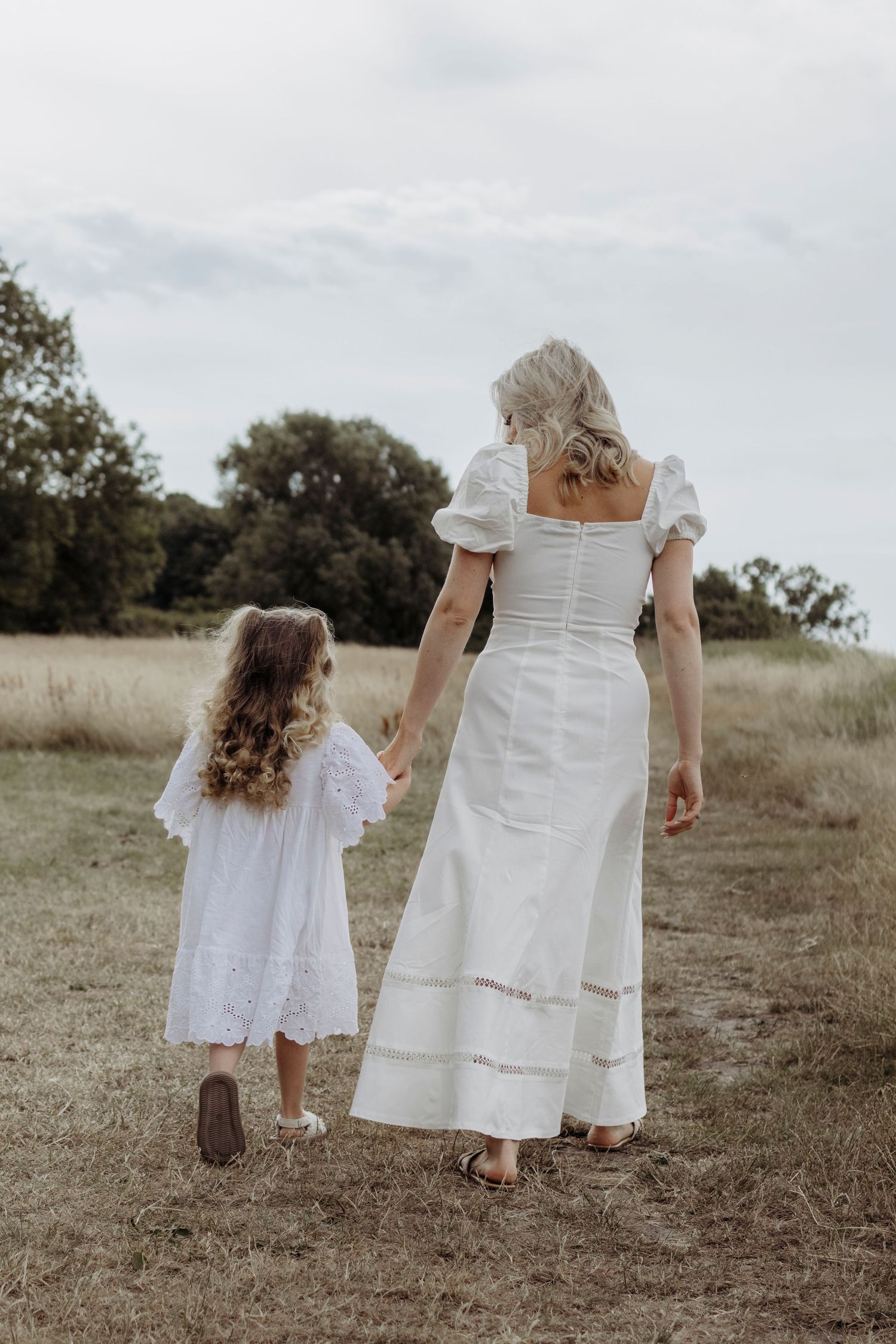 People in white dresses walk together through a grassy hillside on an overcast day.