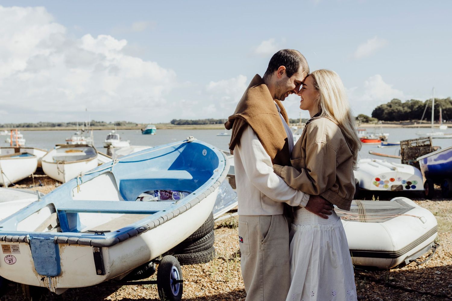 Romantic couple embraces on beach at sunset with boats in background at harbor.