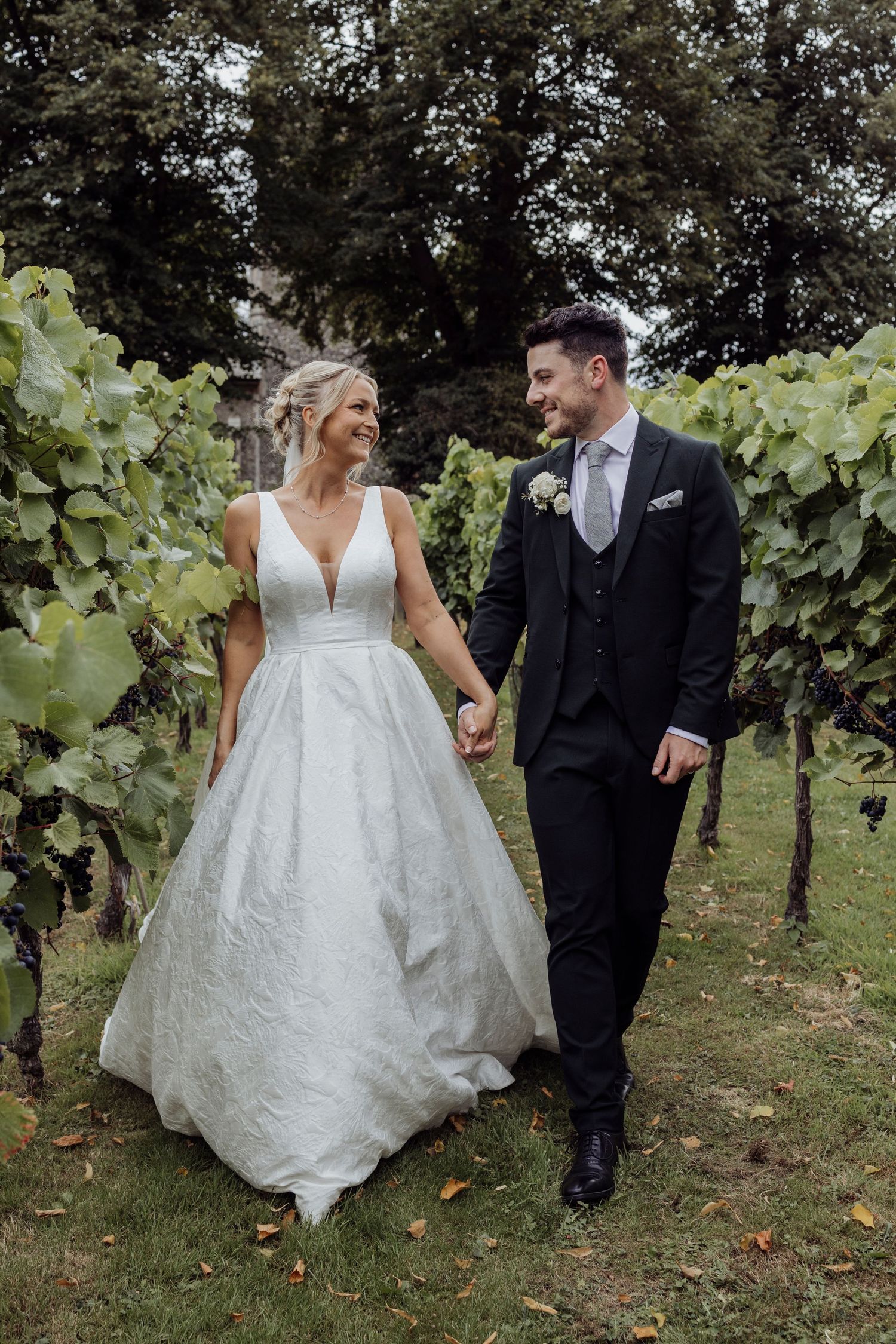 A couple walks through a lush vineyard in elegant wedding attire during autumn.