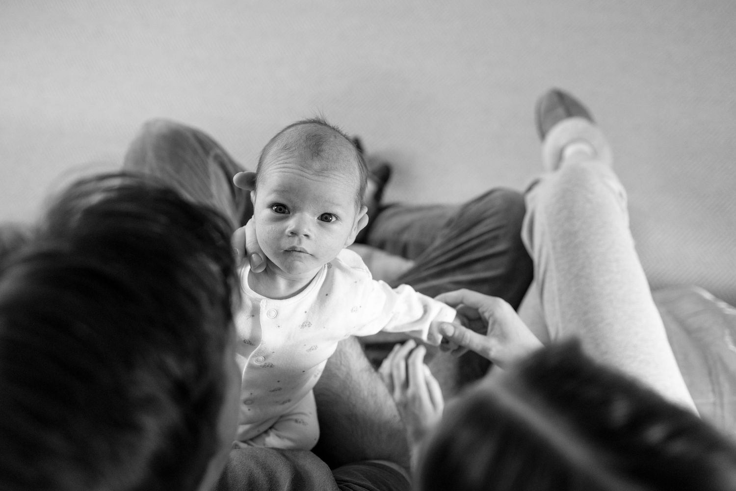 A baby in a white onesie sits with family members on a couch in this black and white photograph.
